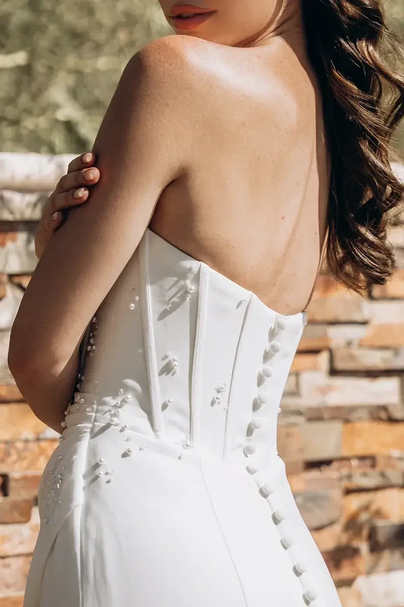 Model is wearing a strapless column wedding dress with a corset-style bodice, sleek satin skirt, and subtle pearl, sequin, and floral details.