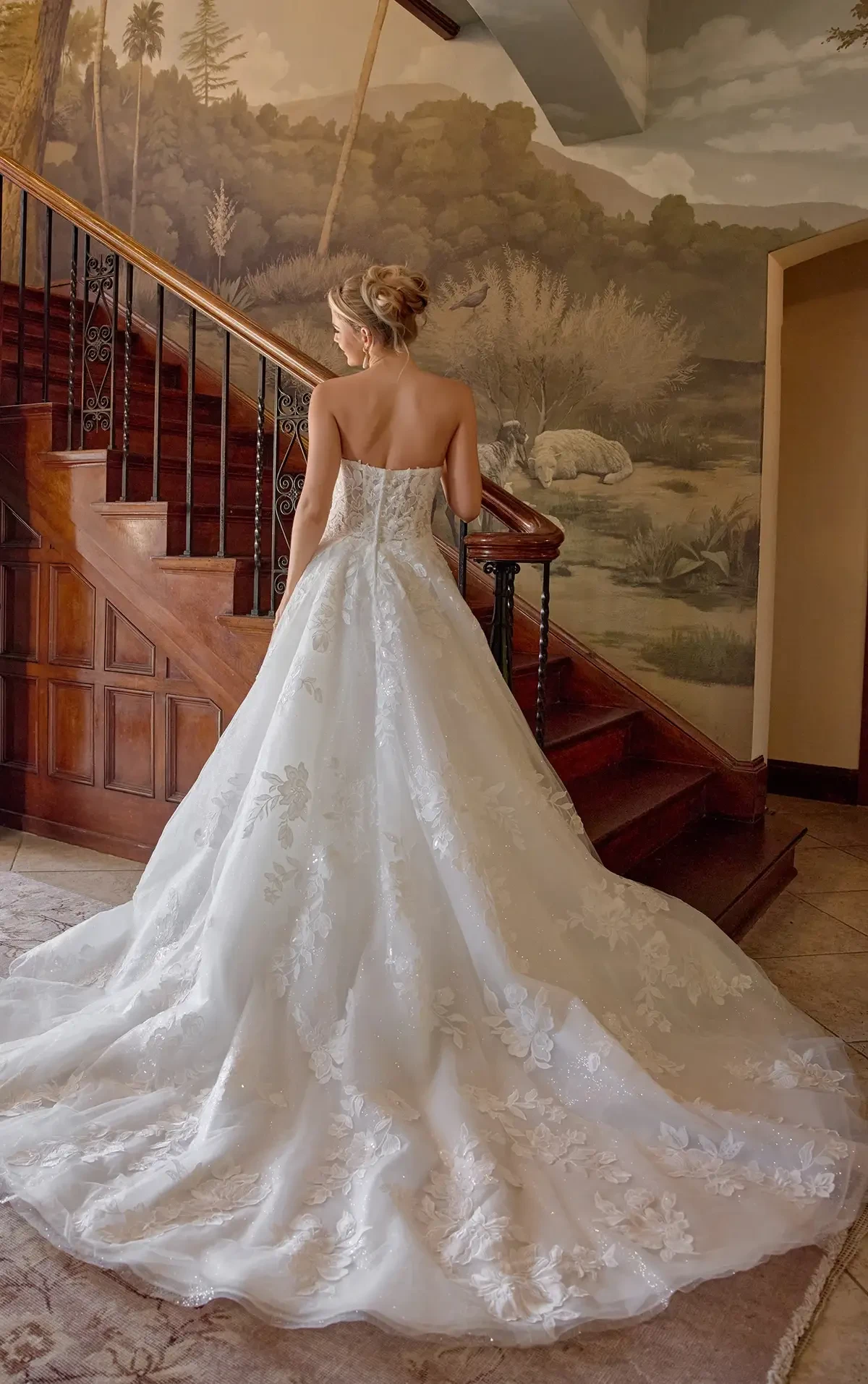 A bride in an elegant white gown with lace detailing stands on a wooden staircase. The background features a mural of a serene landscape.