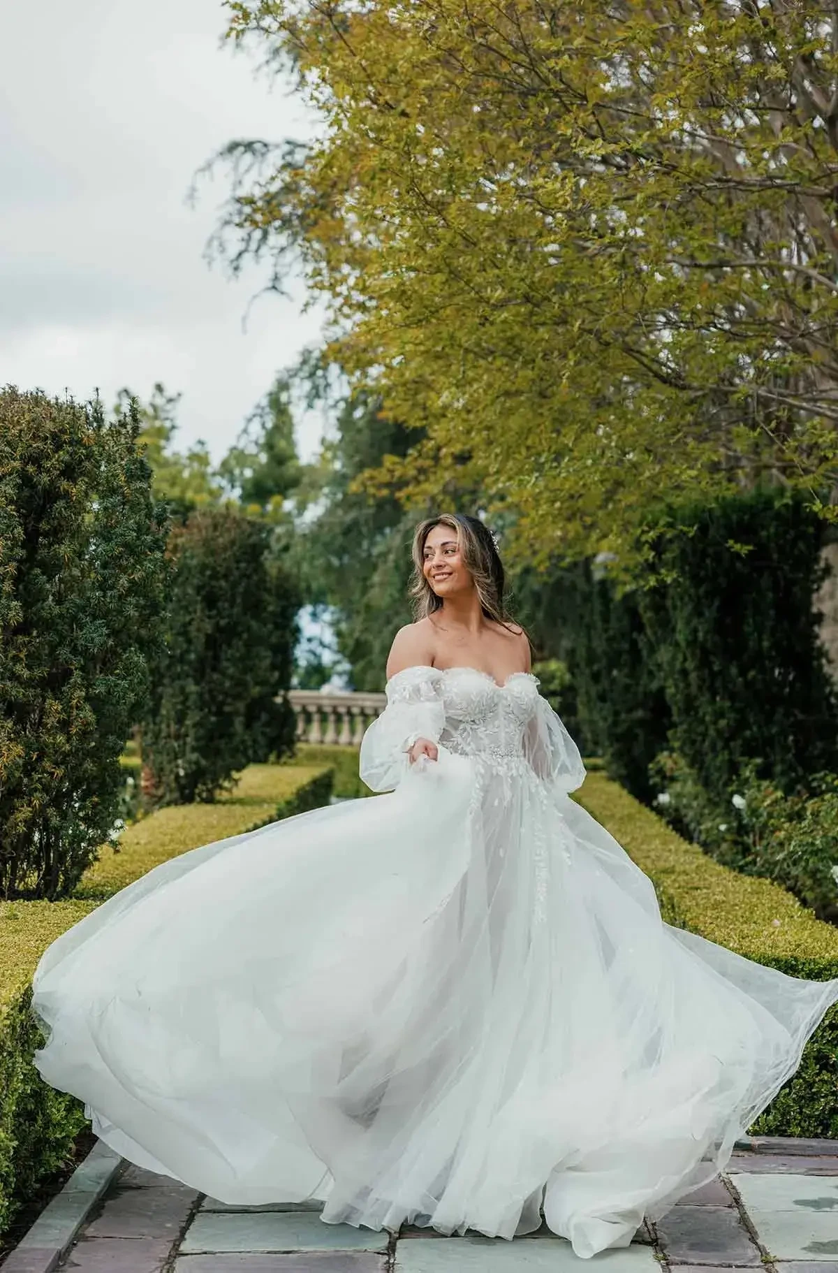 Model is wearing an a-line wedding dress with detachable off the shoulder blouson sleeves and a long train