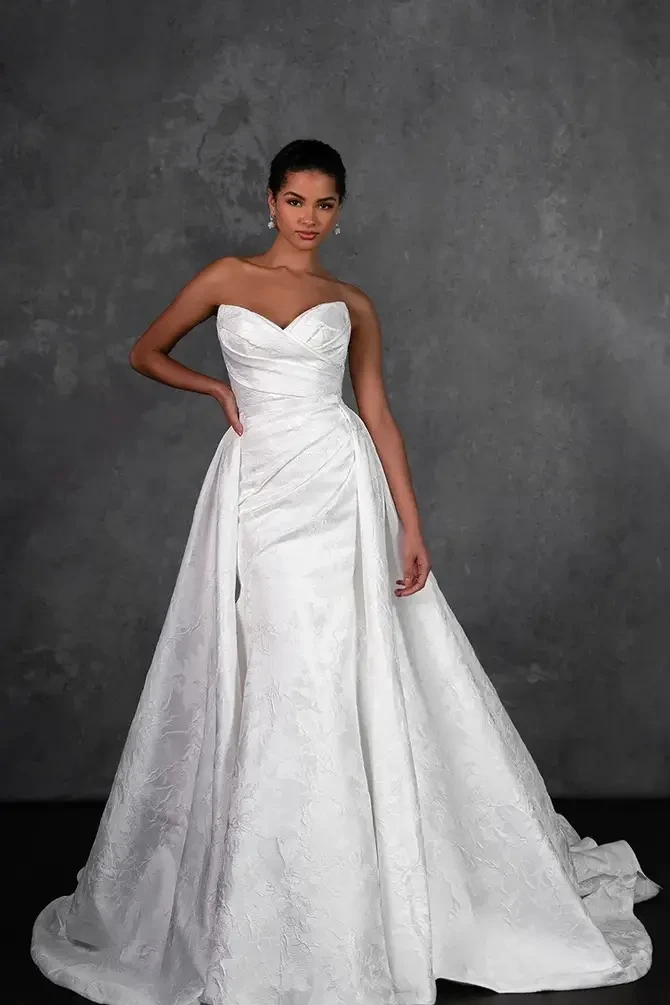 Model is wearing a fit and flare gown with ruched bodice detailing, a gently flared skirt, and a detachable pleated overskirt.
