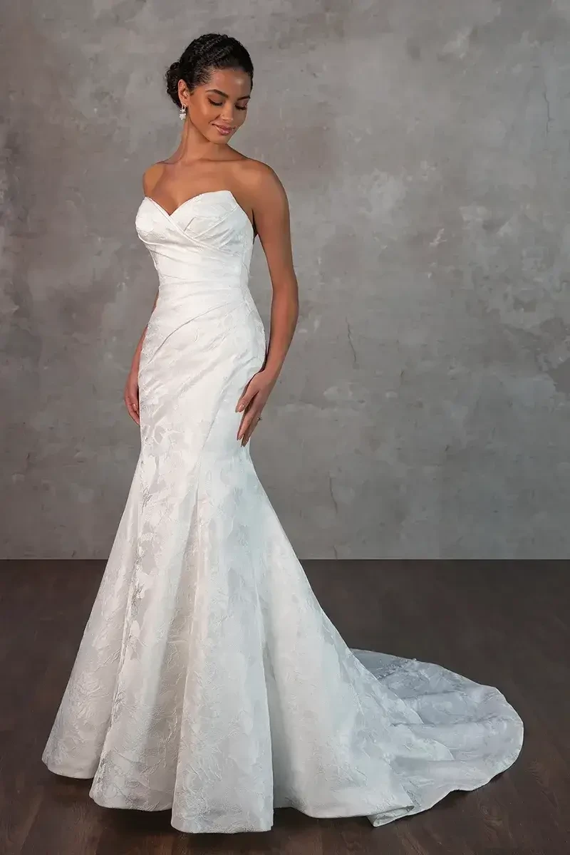 Model is wearing a fit and flare gown with ruched bodice detailing, a gently flared skirt, and a detachable pleated overskirt.
