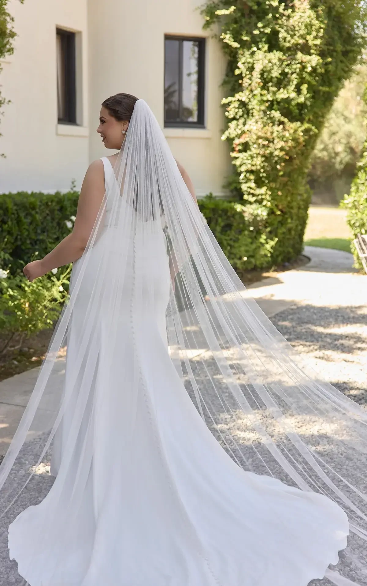 A bride in a flowing white gown and long veil walks on a sunlit path, surrounded by lush greenery and a building in the background, creating a serene and elegant scene.