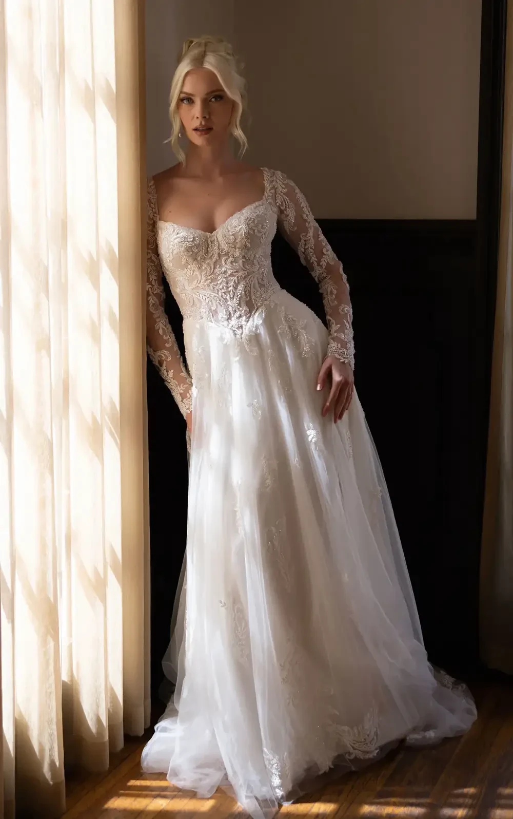 A model wearing a beautiful long-sleeved wedding dress with intricate lace details, standing by a window with soft light illuminating the scene.