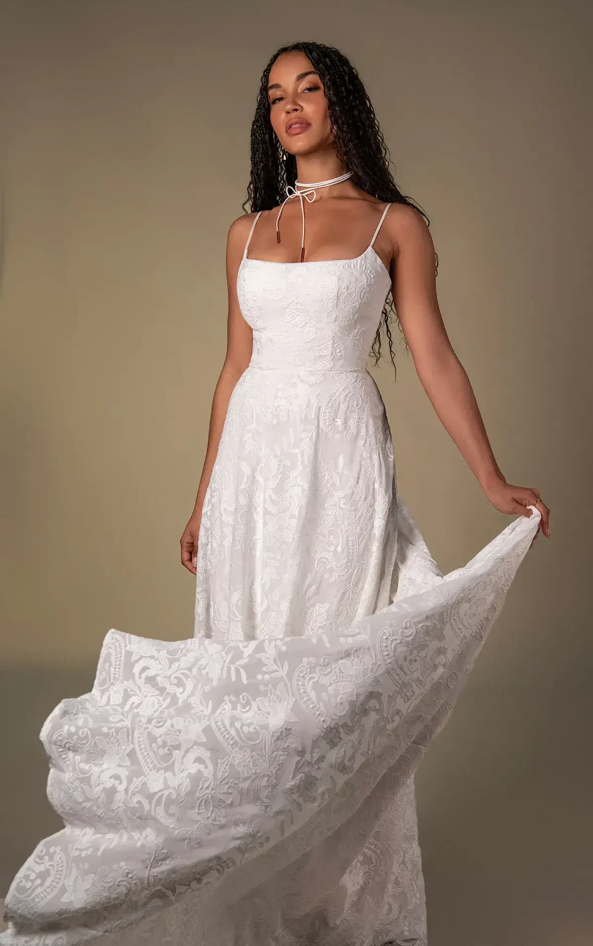 A woman in an elegant white dress with a lace pattern and spaghetti straps poses against a beige background, exuding confidence and grace.