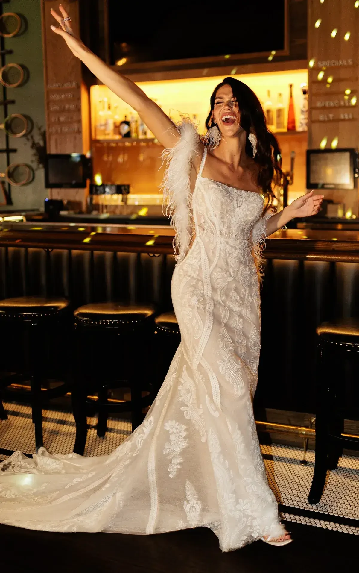 Smiling woman in an elegant white lace gown with feather details, posing joyfully inside a dimly lit bar. She exudes celebration and elegance.