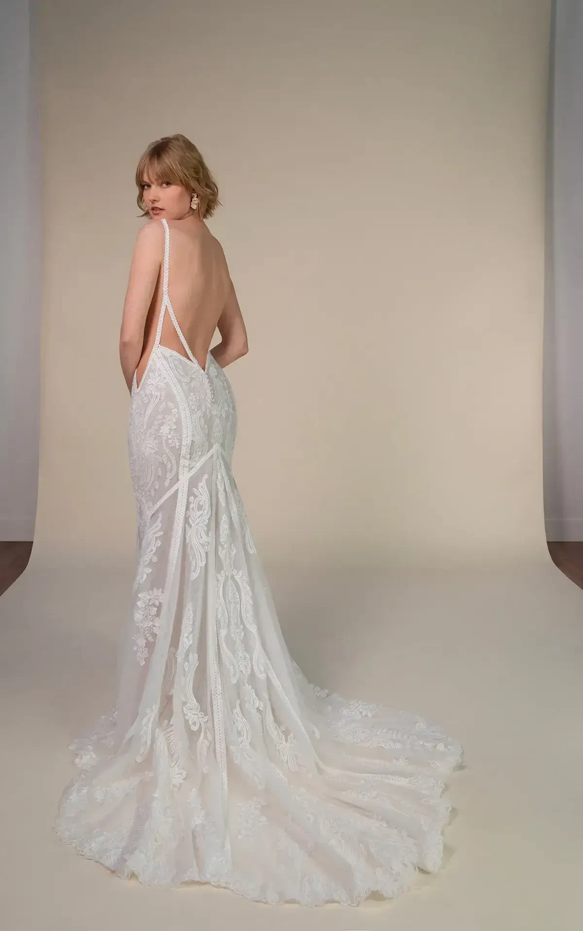 A woman in a backless, lace wedding gown stands on a neutral backdrop. The dress has intricate detailing and a long train. She looks over her shoulder calmly.