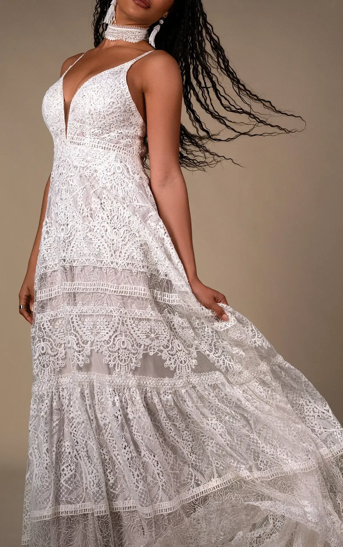 A woman in a flowing white lace dress with intricate patterns stands gracefully, her long hair blowing gently. The mood is elegant and serene.