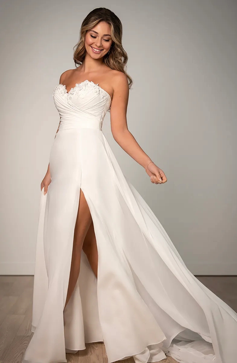 A woman in a strapless white gown smiles gently. The dress has floral lace details and a thigh-high slit, exuding elegance and grace against a simple background.