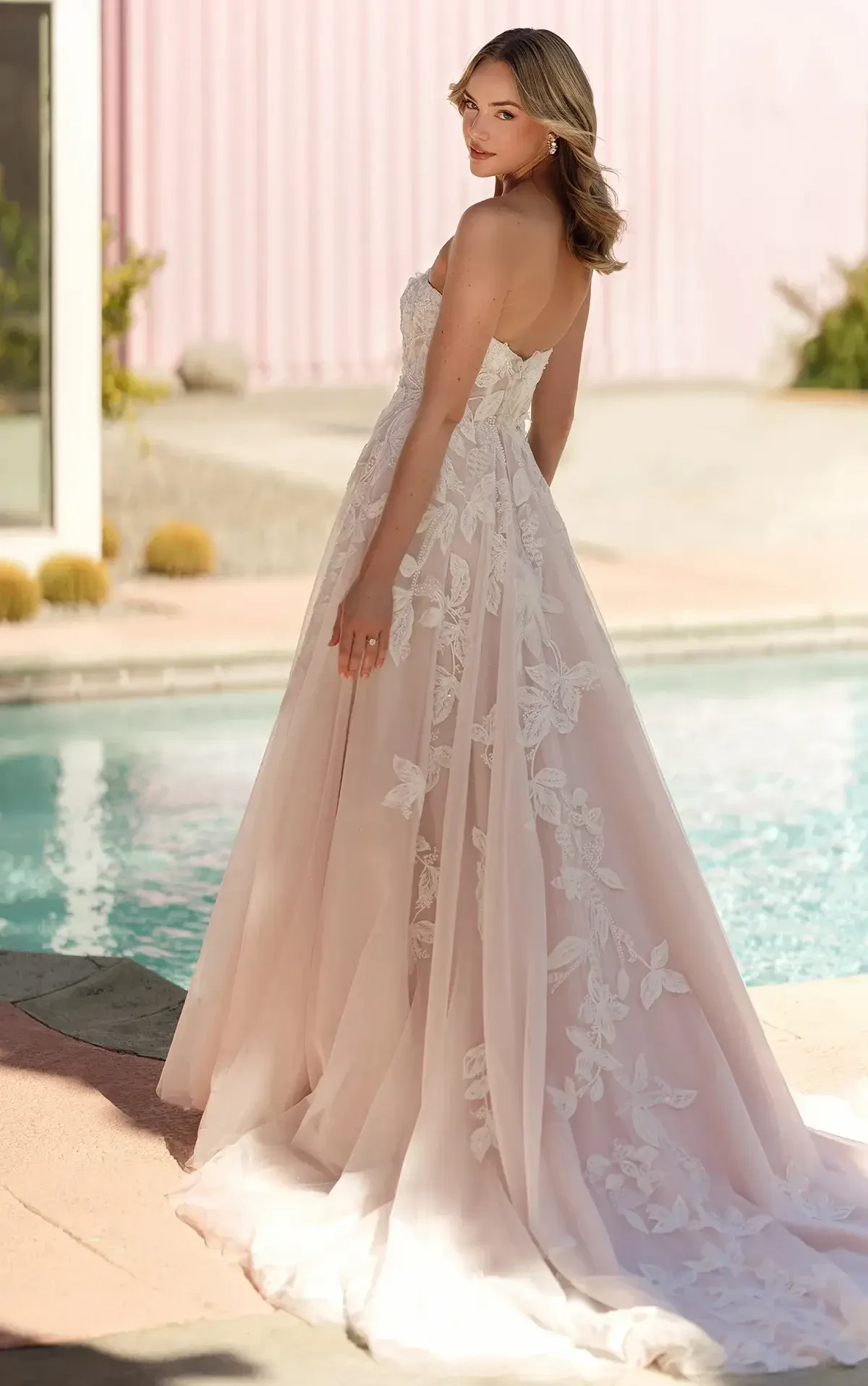 A woman in a flowing strapless wedding gown adorned with lace stands by a sparkling pool. The setting is serene and elegant with a soft pink backdrop.