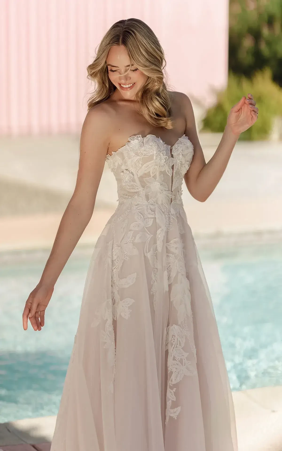 A woman in a strapless lace wedding dress stands by a pool, smiling and looking down. The scene is bright and airy, evoking joy and elegance.