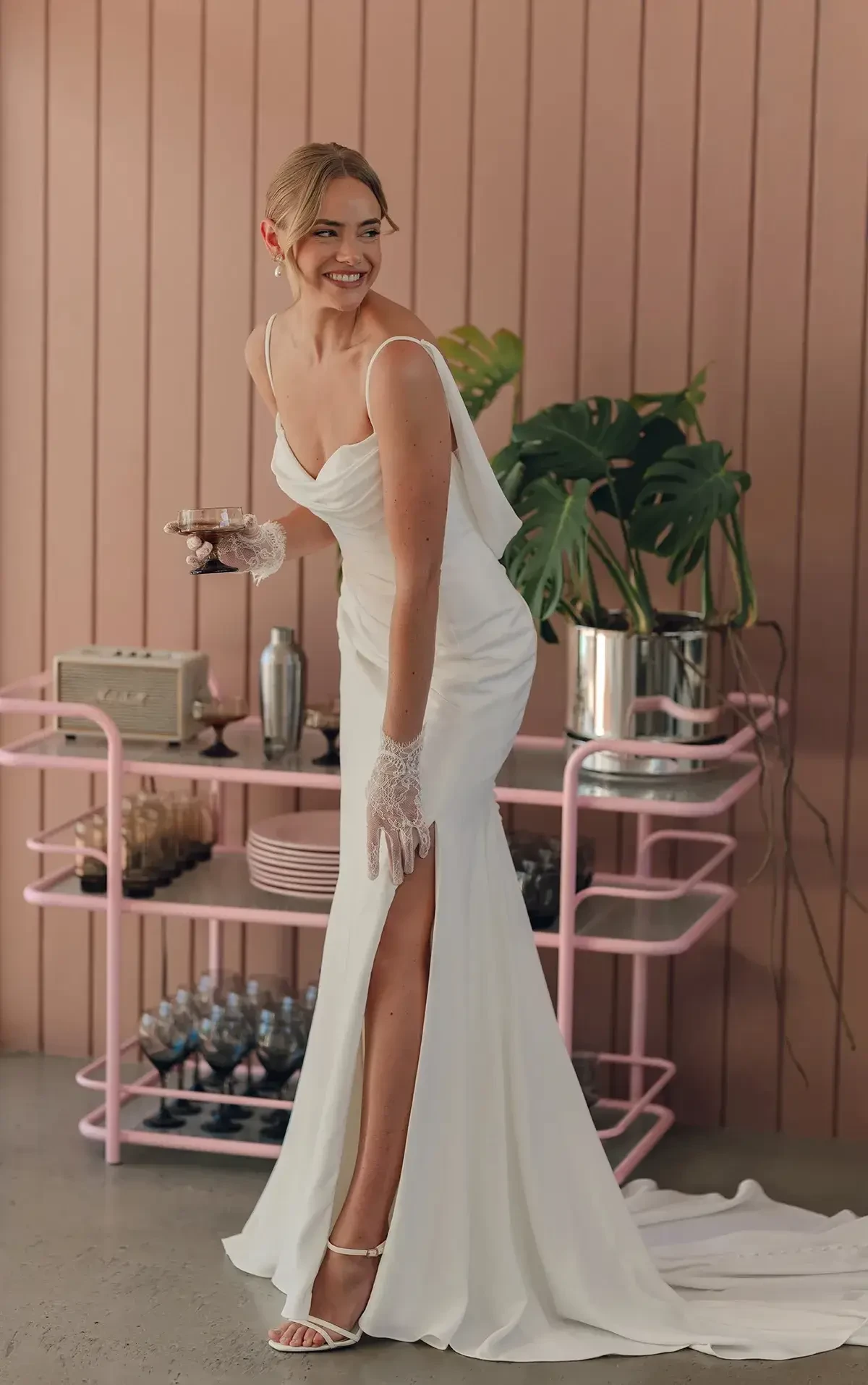 Smiling woman in elegant white gown with lace gloves, holding a cocktail. She's in front of a pink drinks trolley with tropical plants, exuding joy.