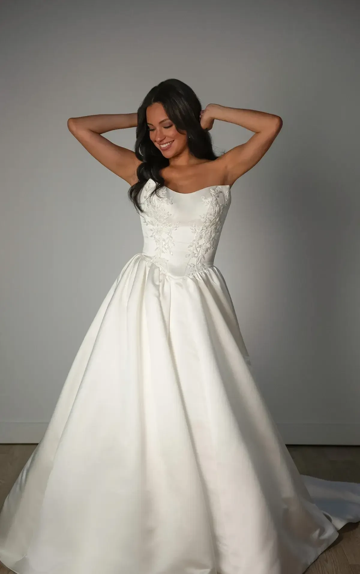 A woman joyfully poses in a strapless, embroidered white wedding dress with a voluminous skirt, set against a neutral gray background.