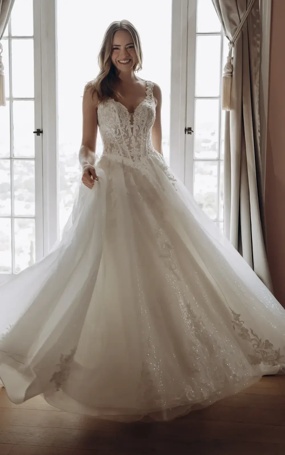 A smiling woman wearing a white wedding dress, standing by a window with light streaming in.