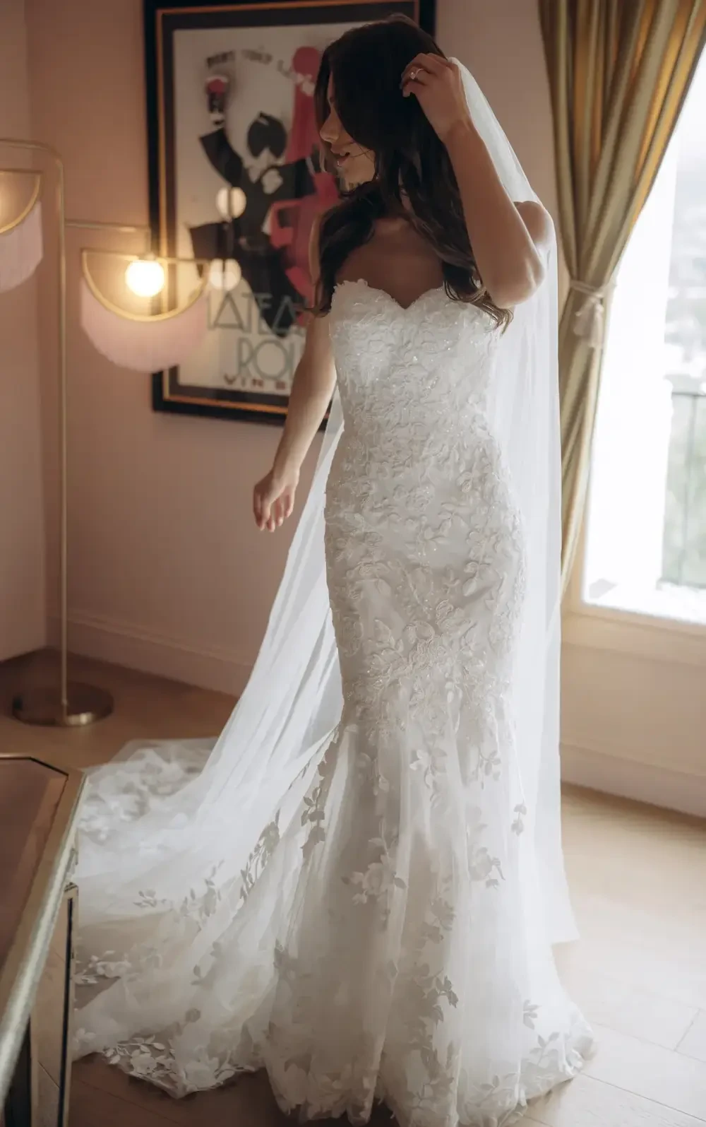 Bride in a stunning lace wedding dress, wearing a veil, standing in a well-lit room with stylish decor.